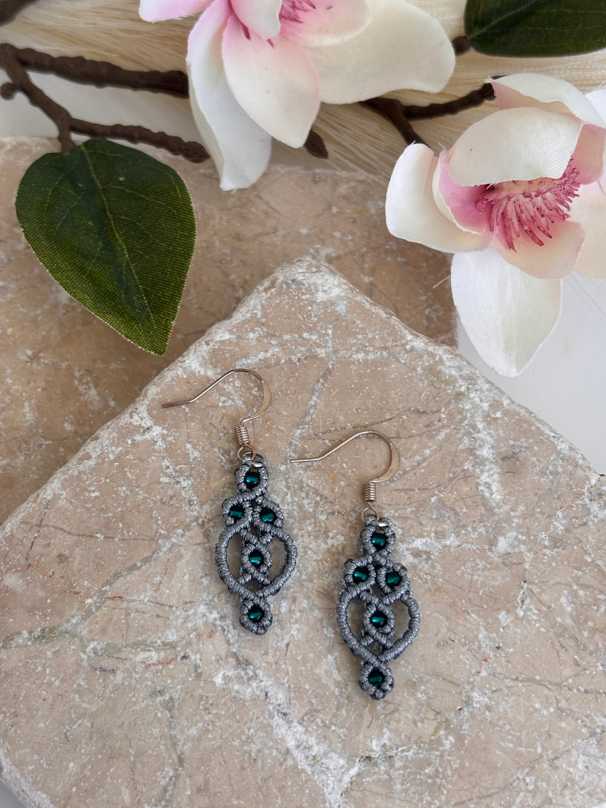 Pair of handmade gray nylon macrame drop earrings with interlocking ring and picot lattice knotwork, 8/0 emerald green seed beads, and silver-tone fish hook wires, displayed on travertine tile with white magnolia flowers and green leaves.