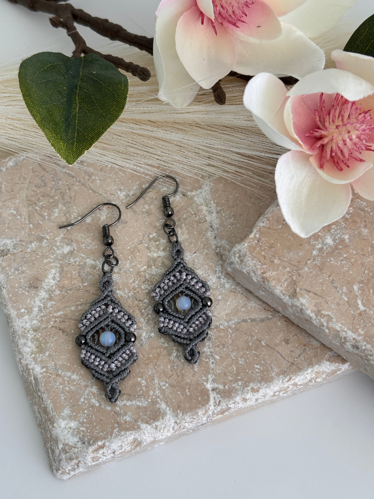 Pair of handmade medium silvery gray nylon macrame drop earrings with open-center diamond silhouette, 4mm round opalite center bead, 3mm gunmetal round metal bead accents, and gunmetal fish hook wires, displayed on travertine tile with magnolia flower and green leaf.