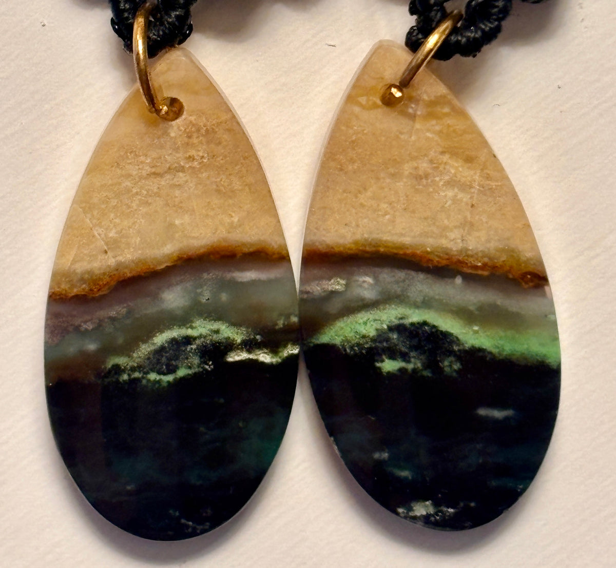 Macro close-up of handmade black macrame drop earrings showing green chalcedony teardrop gemstone detail with natural tan, green, and black mineral banding, and gold-tone jump ring connector.