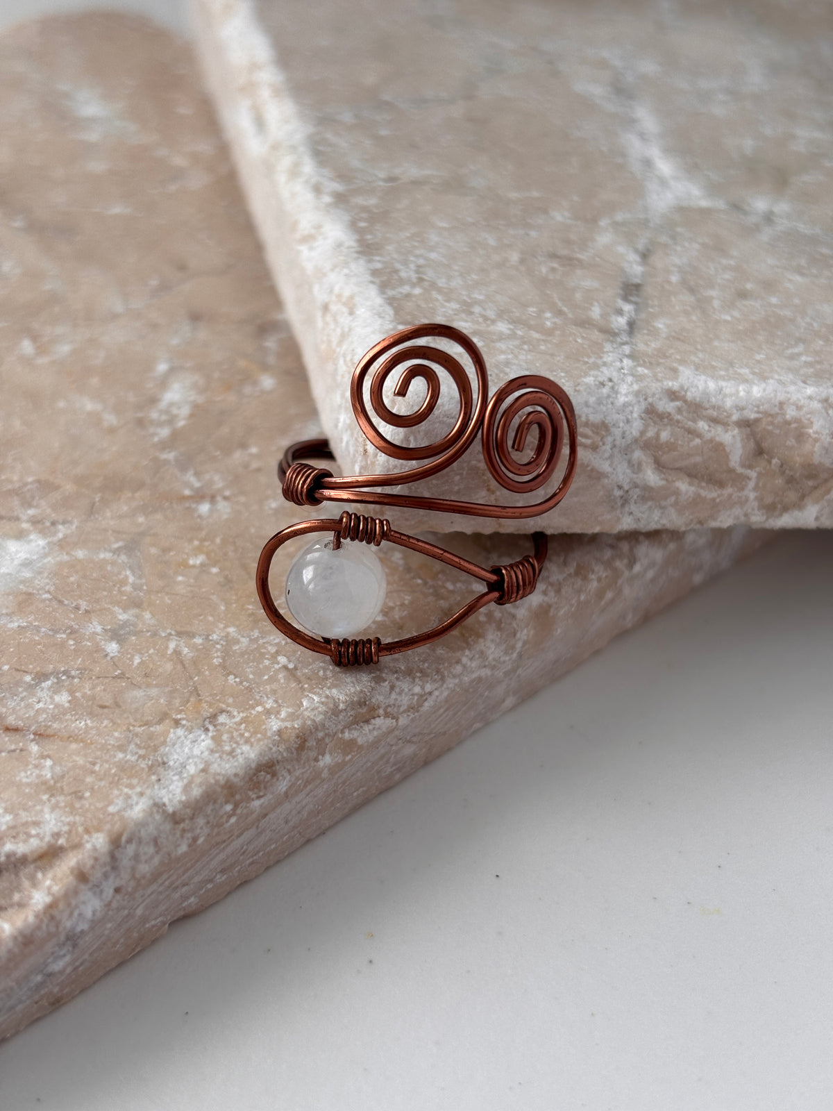 A handcrafted copper wire ring with a double spiral flourish and a wire-wrapped round moonstone bead at the base, leaning against the edge of a natural stone slab against a white background.