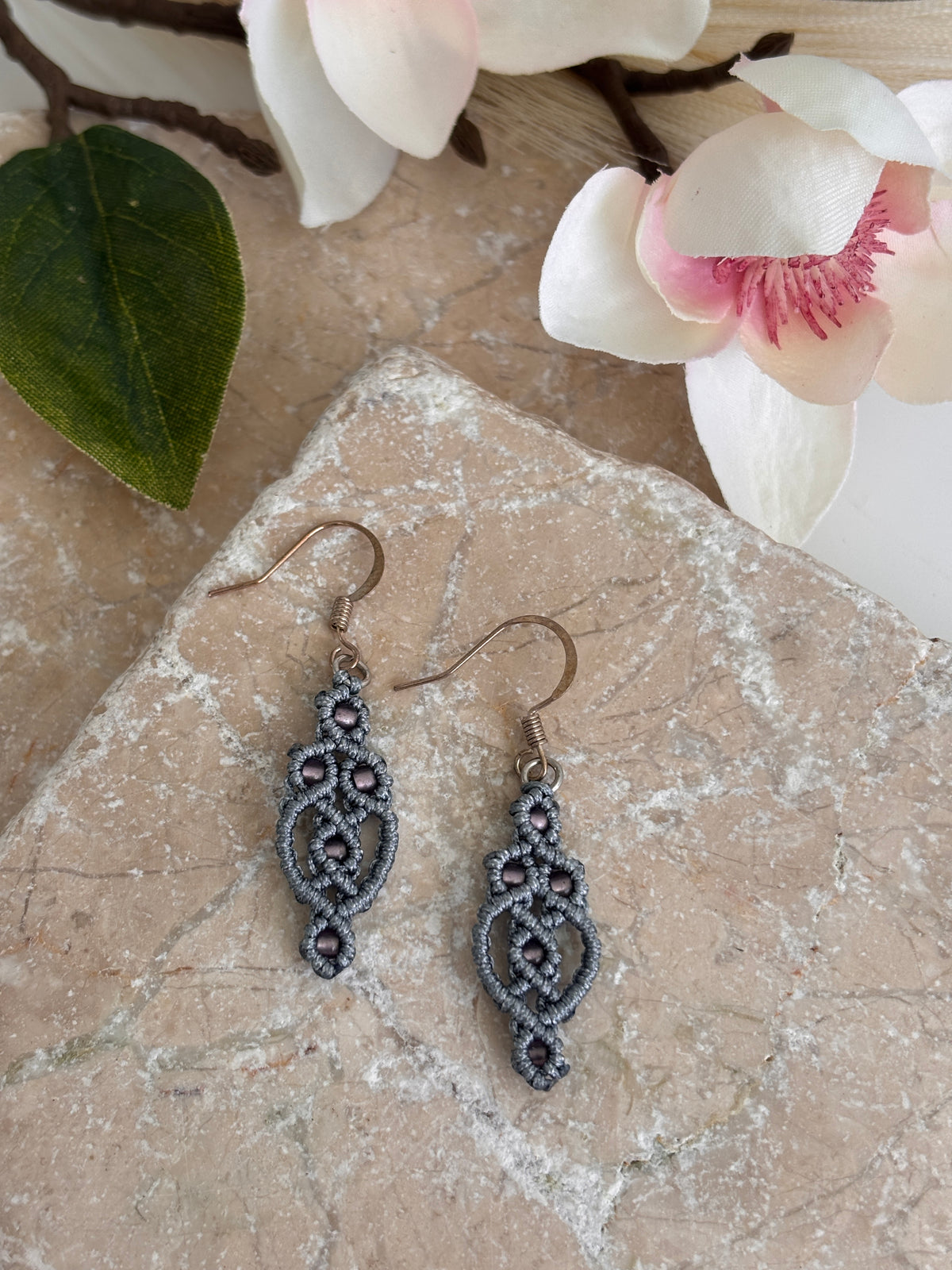 Pair of handmade gunmetal gray nylon macrame lace drop earrings with elongated diamond lattice silhouette and 8/0 plum seed bead accents, displayed on travertine tile with white magnolia flowers and green leaves.