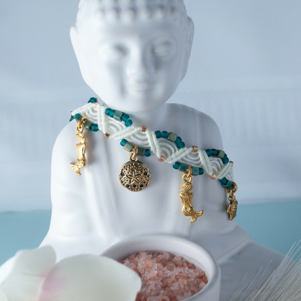 A handmade ivory macrame anklet with teal, gold galvanized, and emerald green seed beads, and gold-toned mermaid, sand dollar, and turtle charms, draped around the neck of a white Buddha statue with a pink salt bowl and soft blue background.