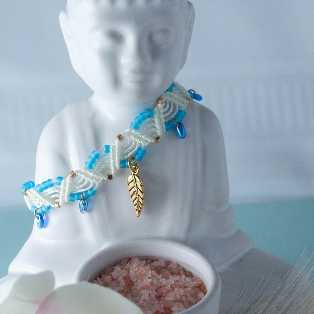 A handmade ivory macrame anklet with turquoise seed beads, iridescent blue Czech glass water drop beads, and a gold-tone leaf charm, draped around a white ceramic Buddha figurine with a small bowl of pink salt crystals in the foreground and a soft blue background.