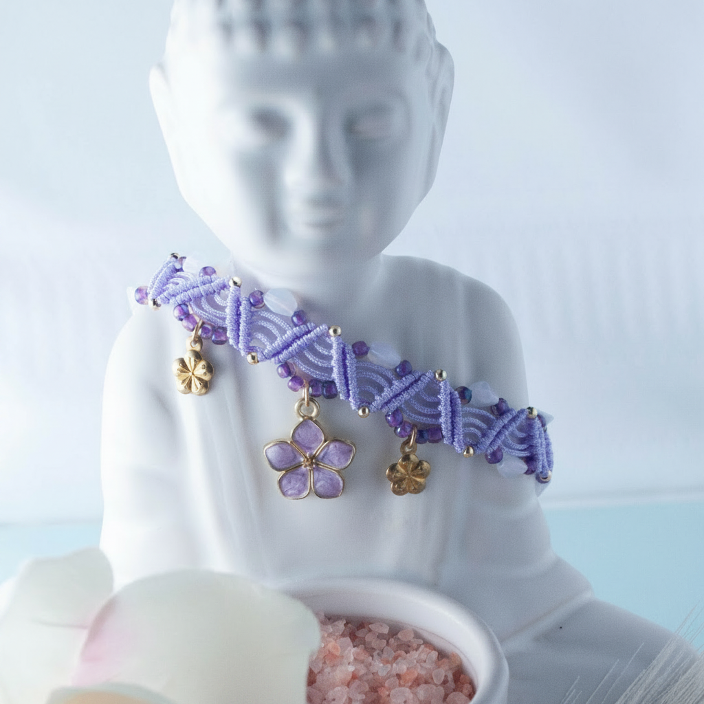 A lavender macrame anklet with purple seed beads, silver accent beads, and three gold flower charms — including a central lavender enamel flower — draped over a white Buddha statue.