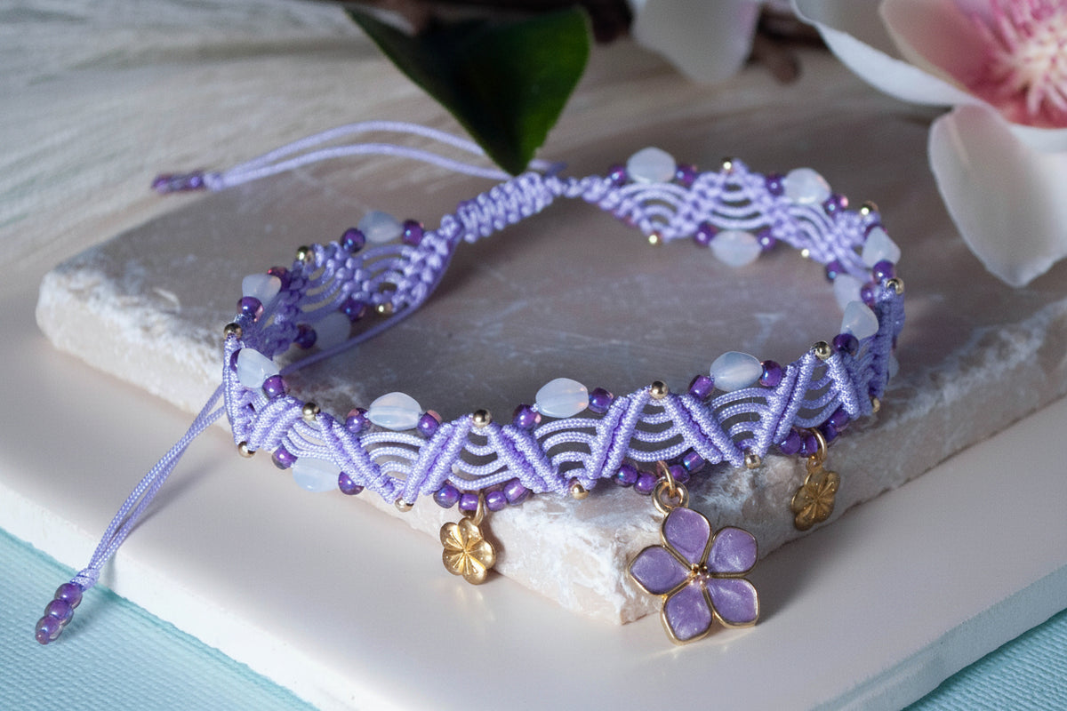 A handmade lavender macrame anklet with purple and moonstone-style beads, gold accent beads, and three gold flower charms including a central lavender enamel flower, displayed on a white stone slab with pink and white florals in the background.