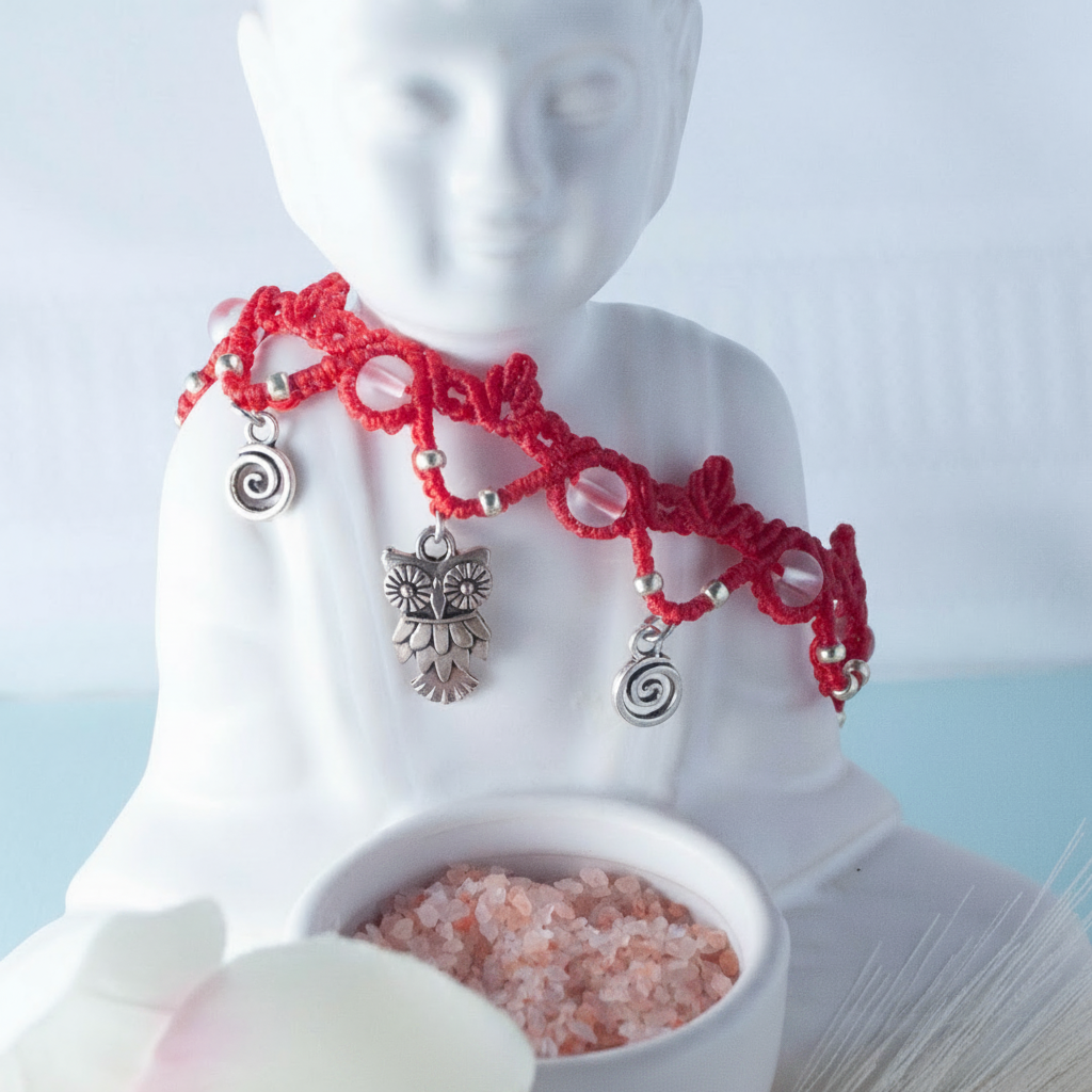 A handmade red macrame anklet with frosted clear mermaid glass beads, silver-tone owl, swirl, and feather charms, draped around a white ceramic Buddha figurine with a small bowl of pink salt crystals in front and a soft blue background.