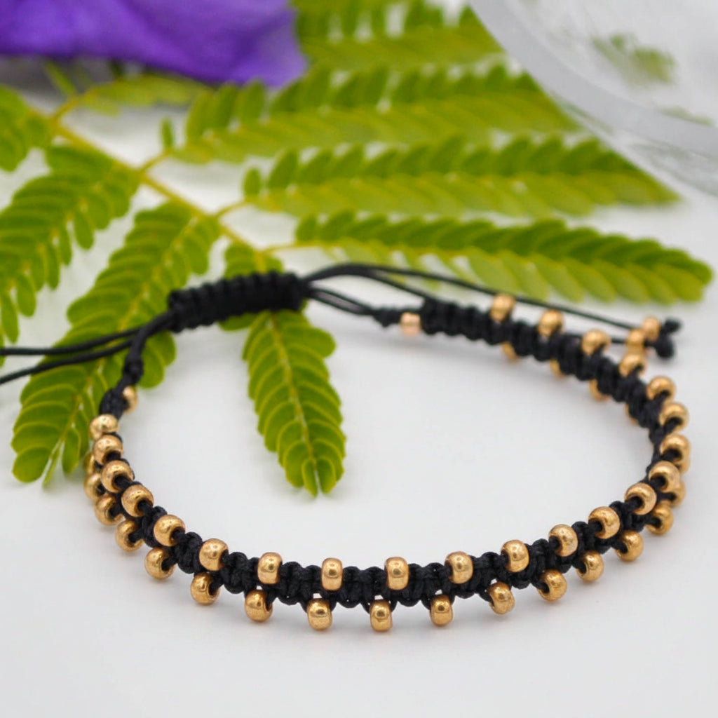 A black macrame bracelet with small galvanized gold seed beads, displayed on a white surface with green leaves and a purple flower in the background.