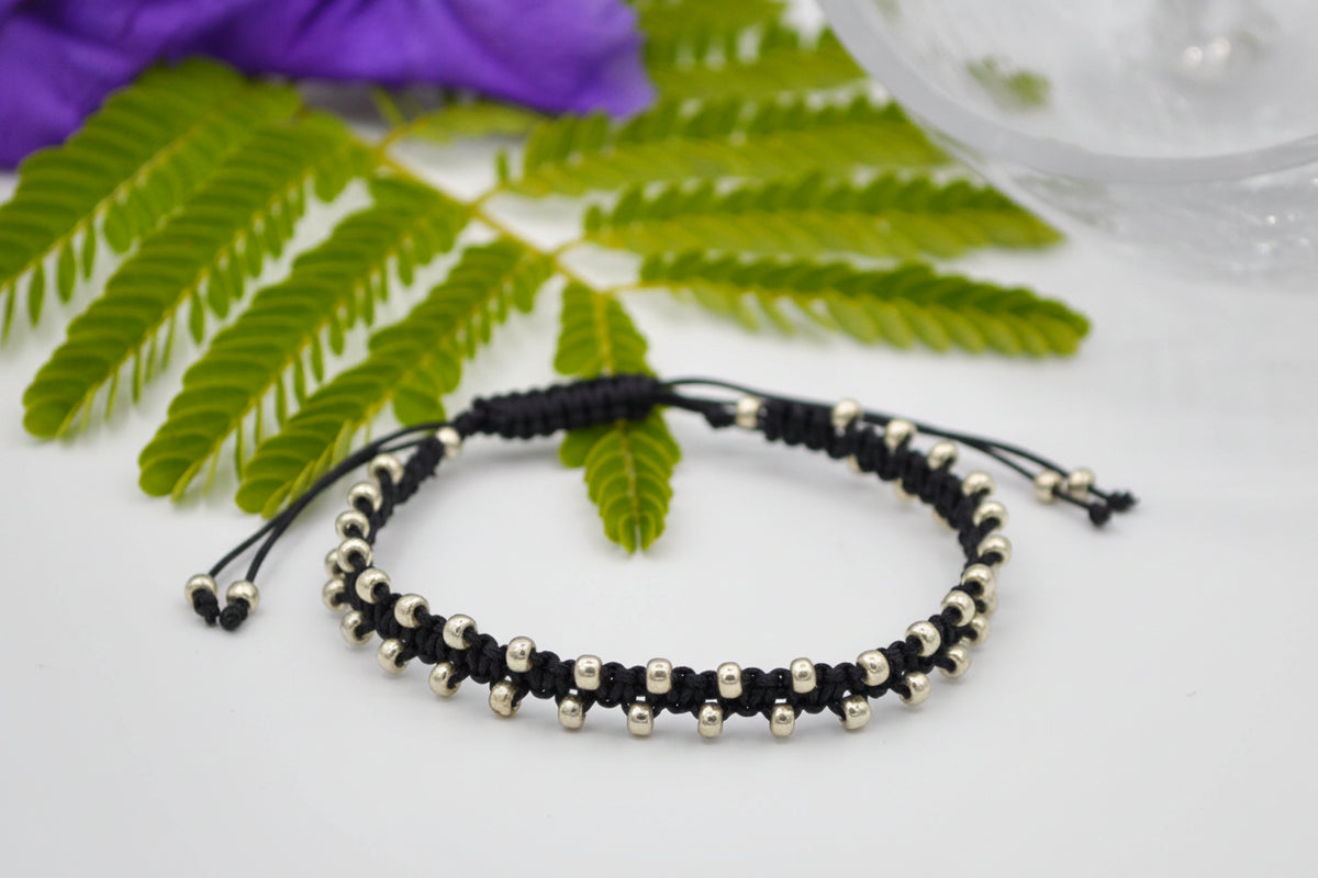 A black macrame adjustable bracelet with galvanized silver seed beads, displayed on a white surface with green leaves and a purple flower in the background.