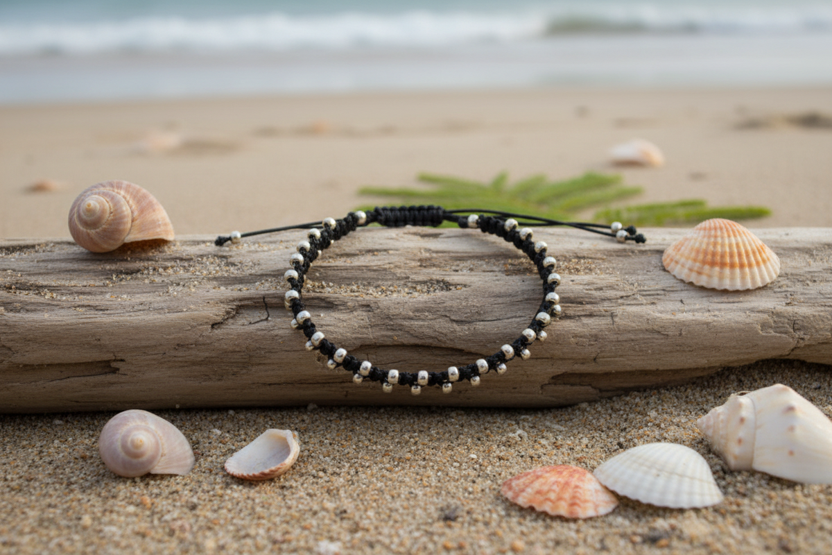A black beaded macrame bracelet with galvanized silver seed beads, styled on a piece of driftwood on a sandy beach with seashells and the ocean in the background.