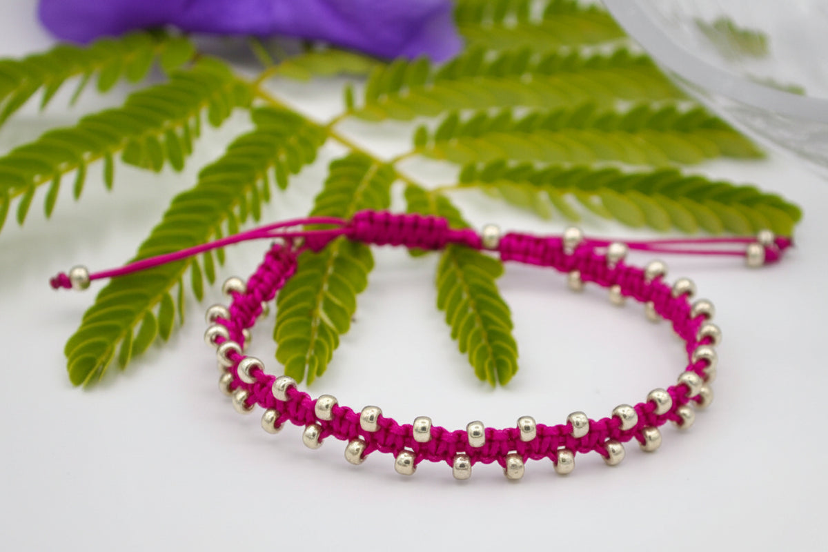 A magenta macrame bracelet with small galvanized silver seed beads, displayed on a white surface with green fern leaves and a purple flower in the background.