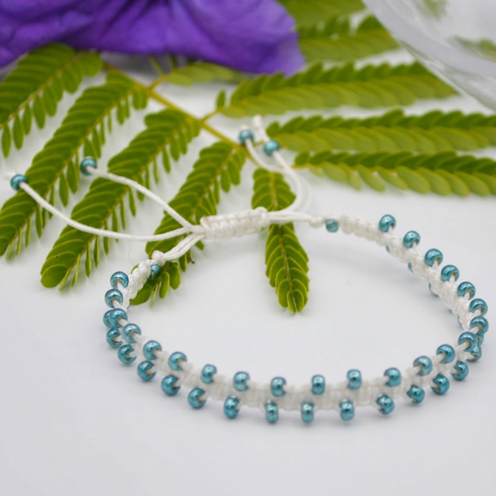 A handmade white macrame bracelet with metallic teal-blue seed beads, styled on a white surface with green leaves and a purple flower in the background.