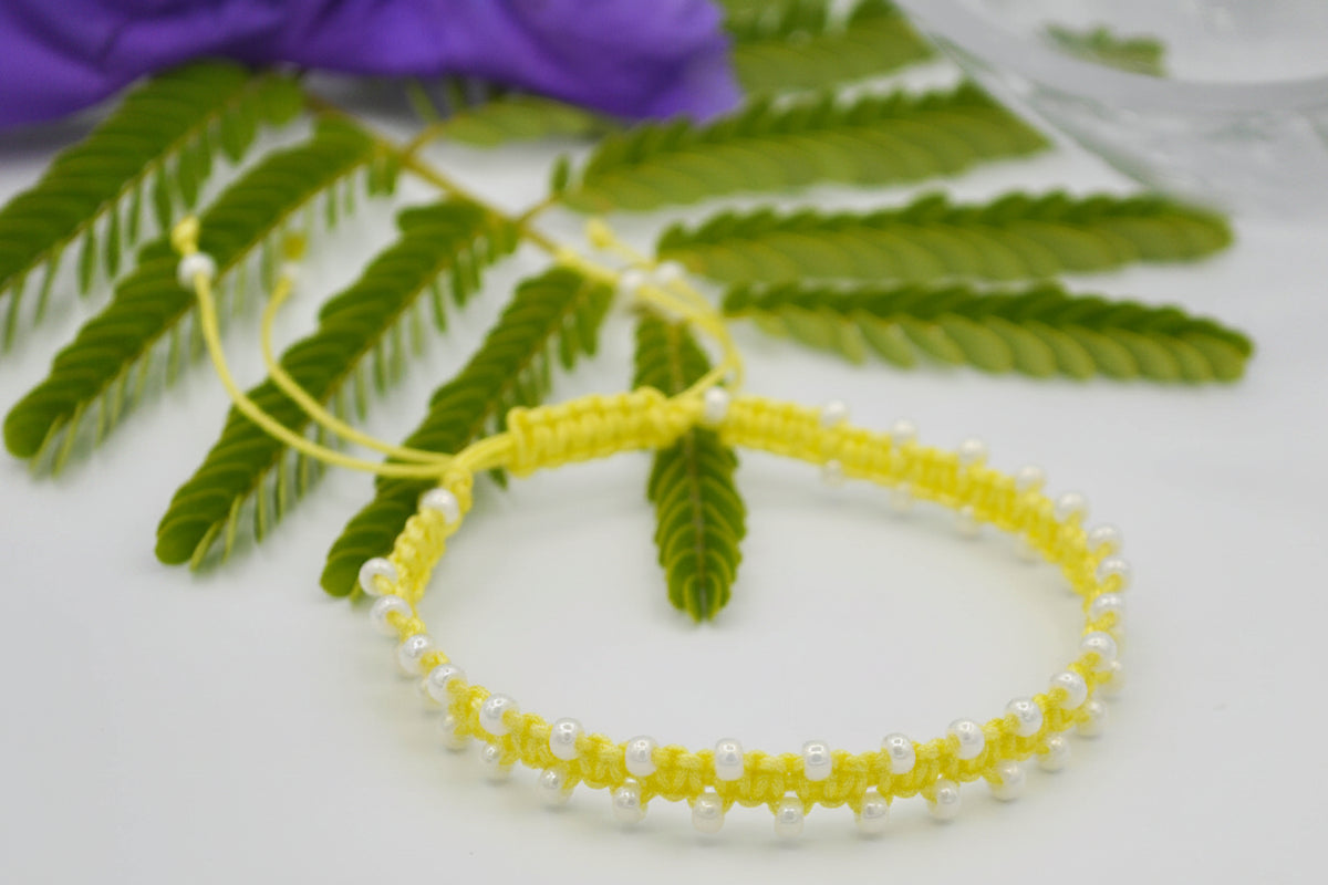 A yellow macrame bracelet decorated with small pearlescent white seed beads, displayed on a white surface with green leaves and a purple flower in the background.