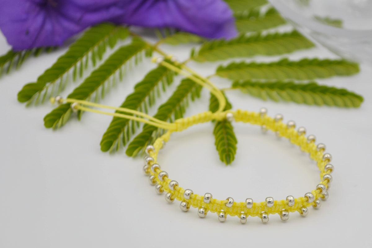 A pale yellow macrame bracelet with small galvanized silver seed beads, displayed on a white surface with green leaves and a purple flower in the background.