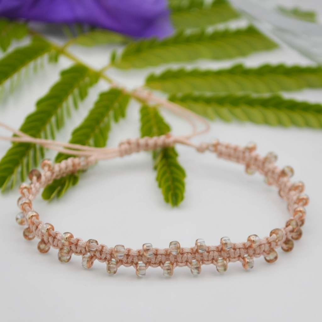 A handmade beaded macrame bracelet with a light peach woven cord and clear, copper edged seed beads, displayed on a white surface with green leaves and a purple flower in the background.