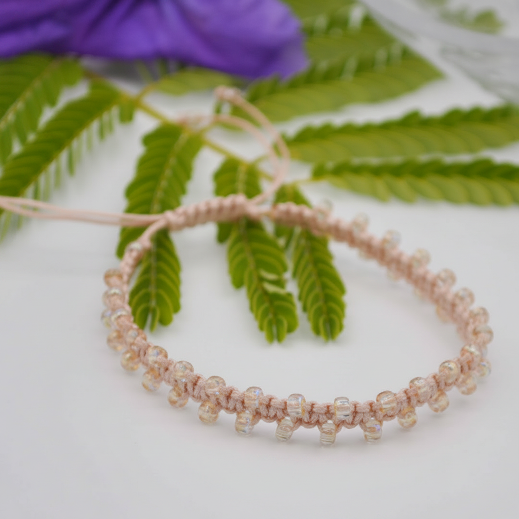 A handmade macrame bracelet in a light peach color, featuring small iridescent yellow seed beads and a knotted cord closure, displayed on a white surface with green leaves and a purple flower in the background.