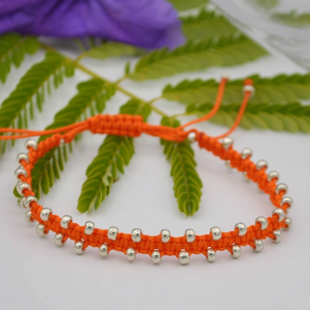 An orange macrame bracelet with small galvanized silver seed beads, displayed on a white surface with green leaves and a purple flower in the background.