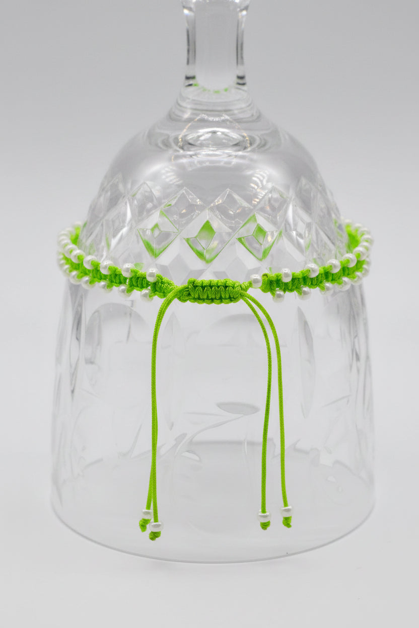 A lime green macrame bracelet or anklet with pearlescent white seed beads, and hanging cords ends, displayed on a clear cut-glass tumbler against a light gray background.
