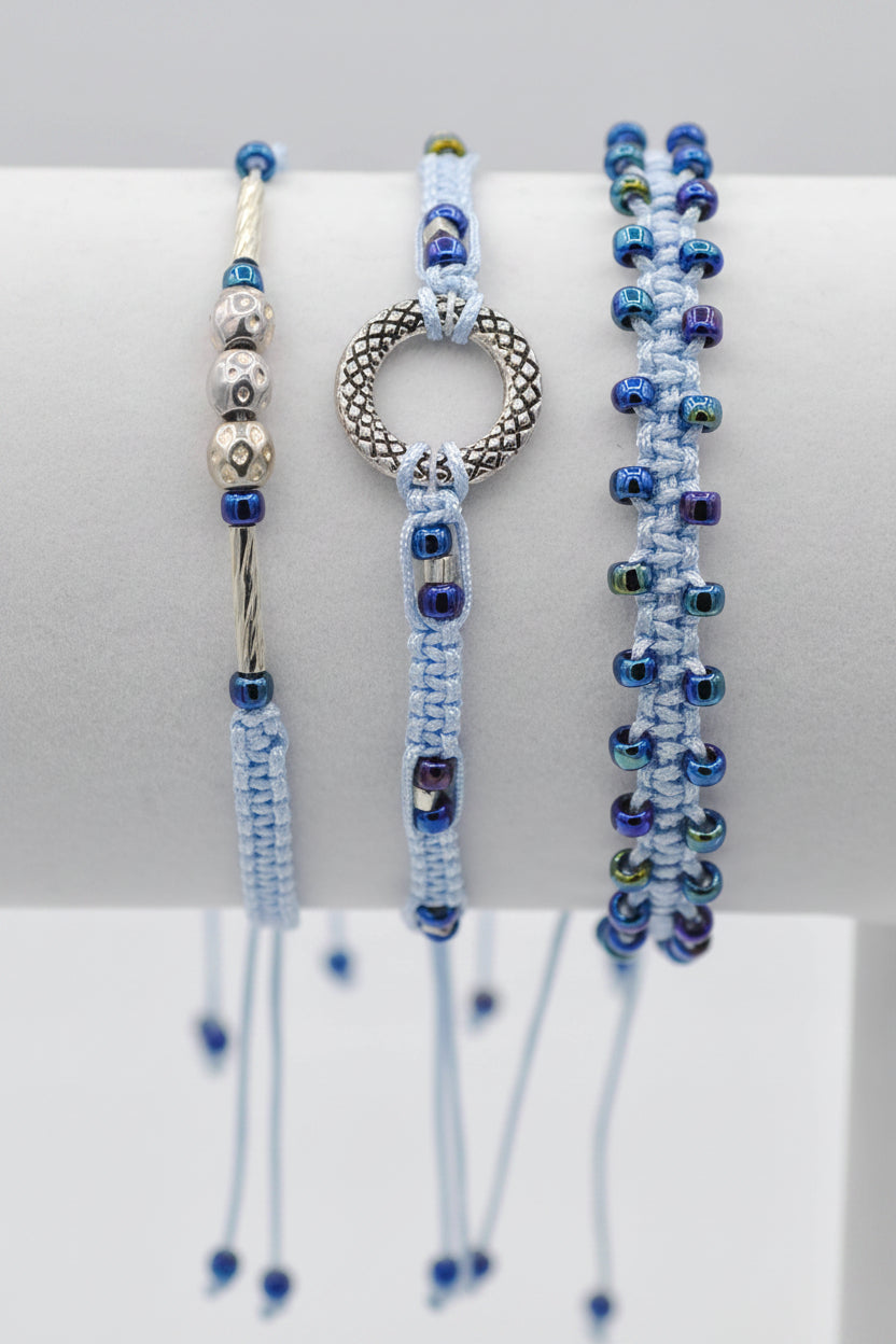 Three handmade macrame bracelets displayed on a white cylindrical stand. The bracelets feature dark metallic blue seed beads and silver-toned metal accent beads, including a textured circular ring,