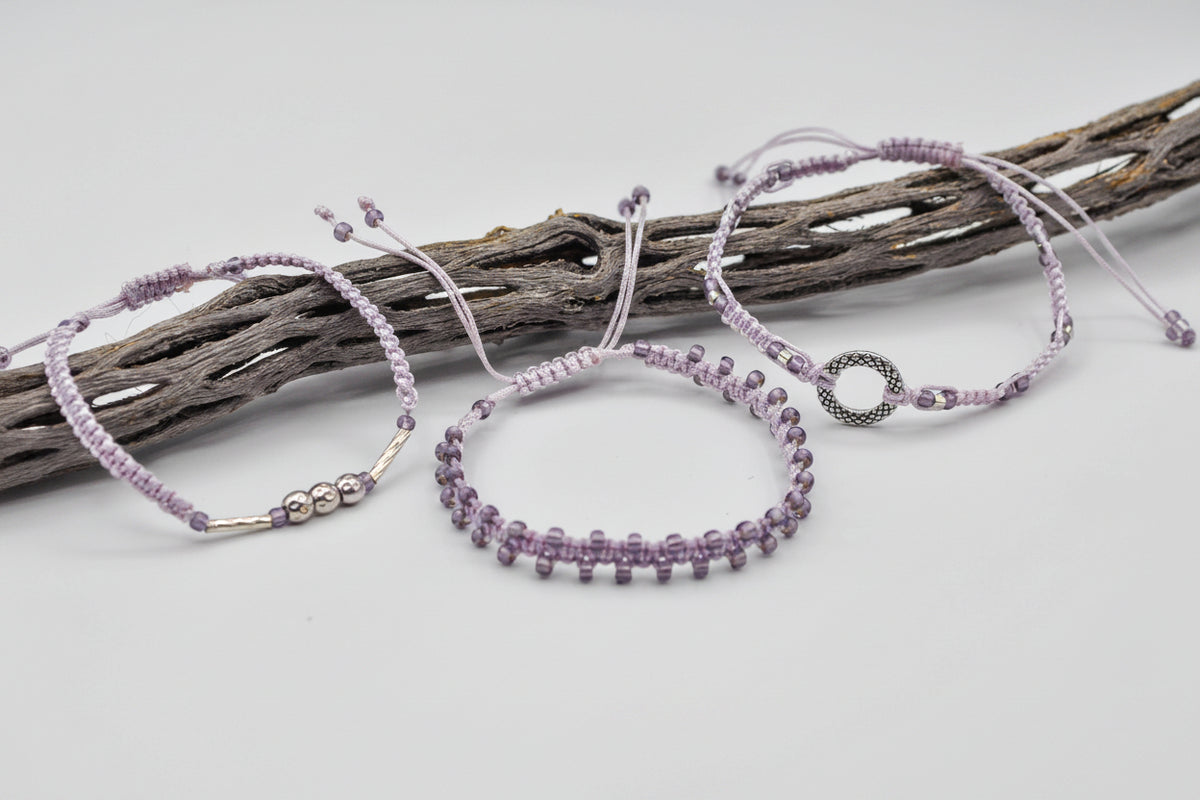 Three light purple macrame bracelets with lavender seed beads and silver-tone metal accent beads in various shapes arranged on a piece of weathered driftwood against a light gray background.