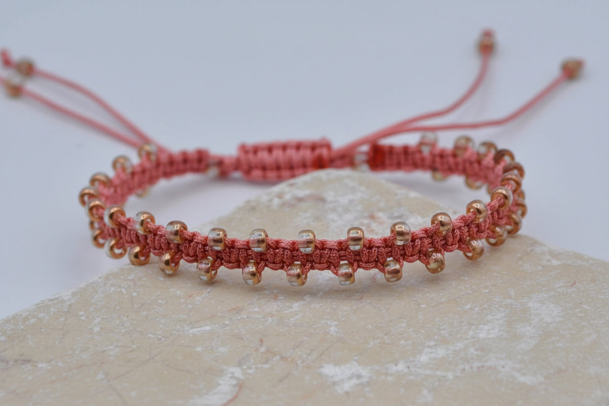 A coral macrame bracelet with small clear, copper-toned seed beads, displayed on a light stone surface against a white background.