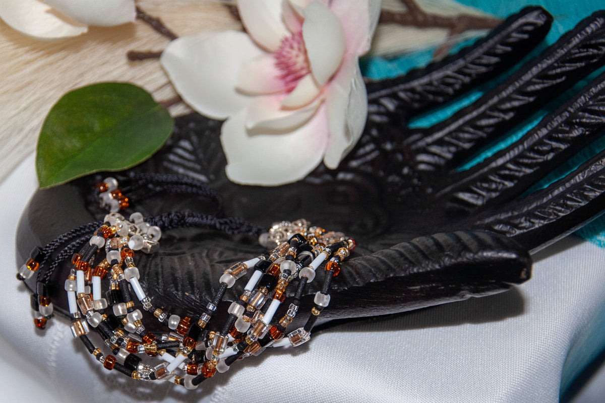 A handmade seven strand seed bead bracelet in black nylon macrame cord with mixed frosted white, matte black, amber rootbeer, and clear glass seed beads, cube beads, and bugle beads, draped over a carved black decorative hand prop with a pink magnolia flower and green leaf in the background
