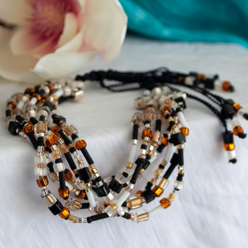 Seven-strand black macramé bracelet with mixed white, black, amber, and clear beads styled on white fabric beside a pink flower