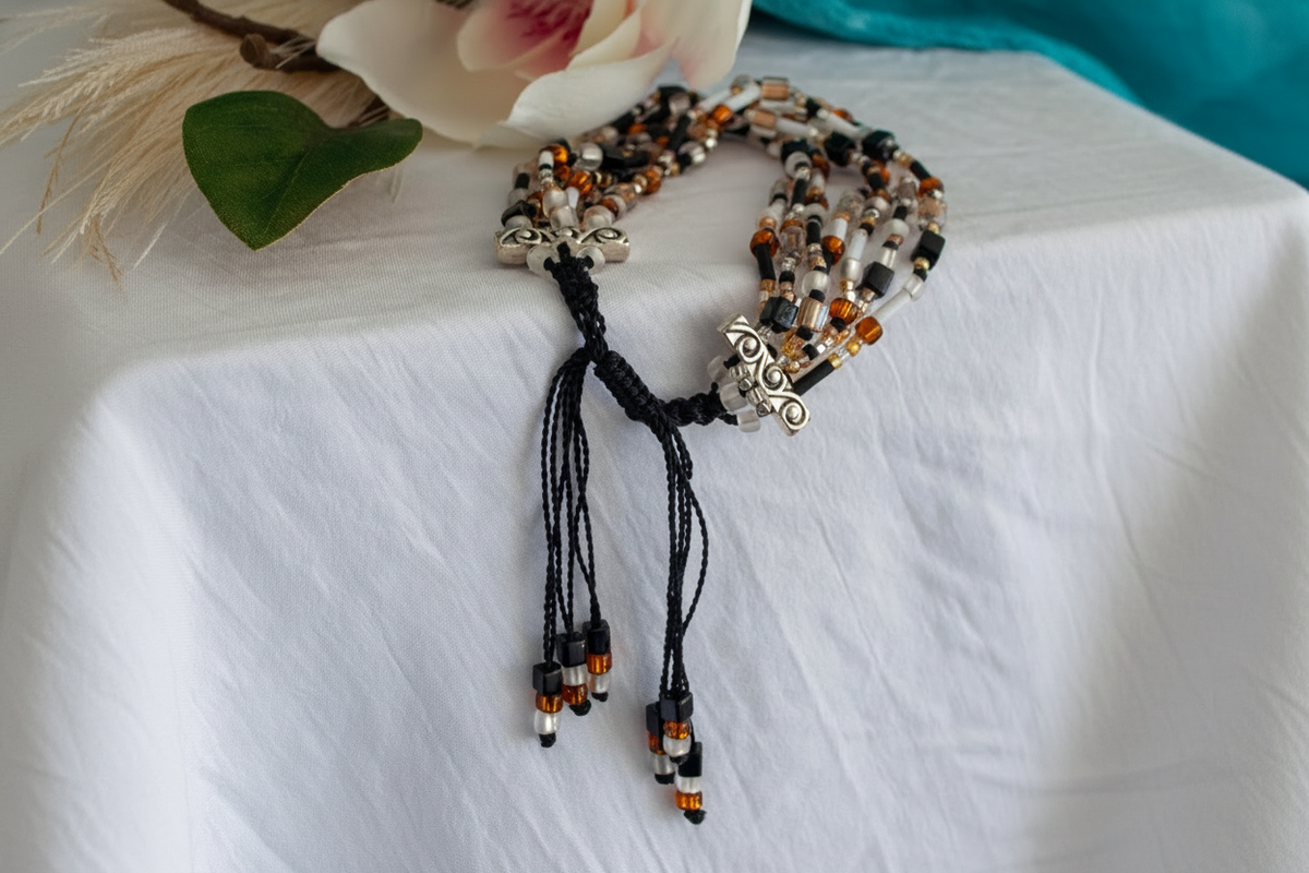 Seven-strand seed bead bracelet in black nylon macramé cord with frosted white, matte black, amber rootbeer, and clear beads, shown with its adjustable slide closure on white linen