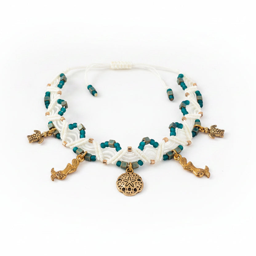 A handmade ivory macrame double zig-zag anklet with teal, gold galvanized, and matte emerald green seed beads, and gold-toned mermaid, sand dollar, and turtle charms, displayed on a white background showing the full piece and slide closure.