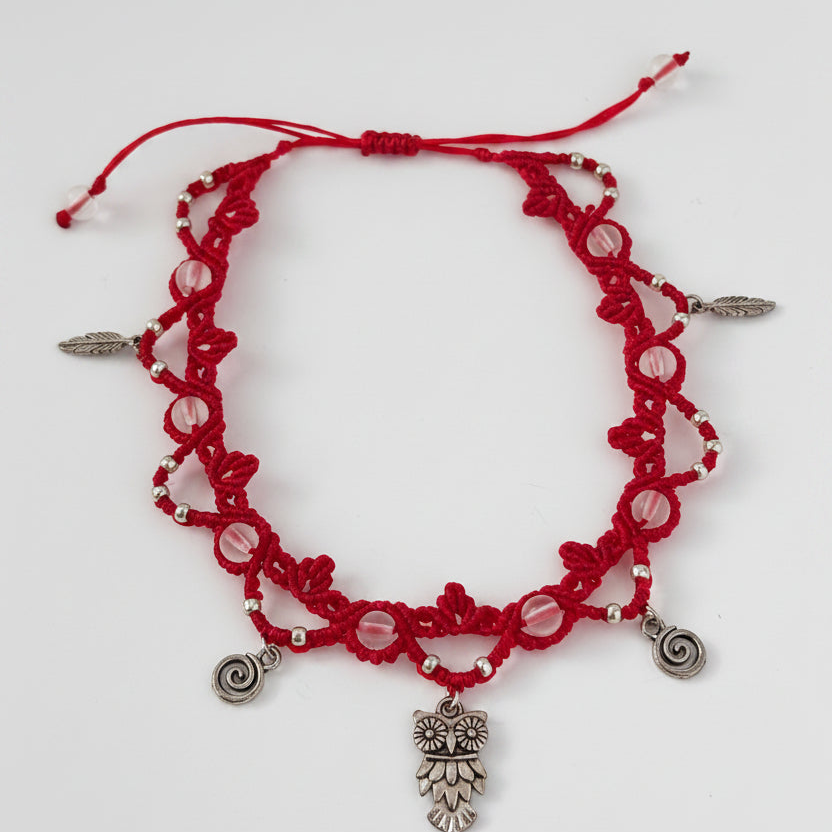 A red macrame beaded anklet with frosted clear mermaid glass beads, silver-toned charms, including an owl pendant, spiral charms, and small feather accents, displayed on a plain white background.