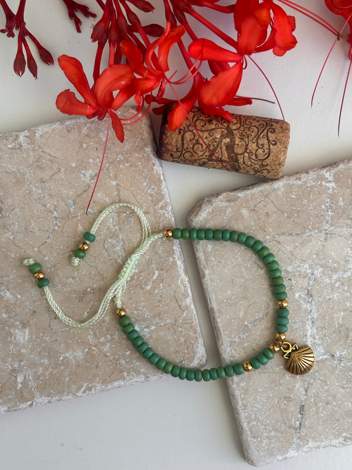 A beaded bracelet with 6/0 green round beads, a mint green adjustable cord, and a brass shell charm, styled on stone tiles with red flowers and a cork in the background.