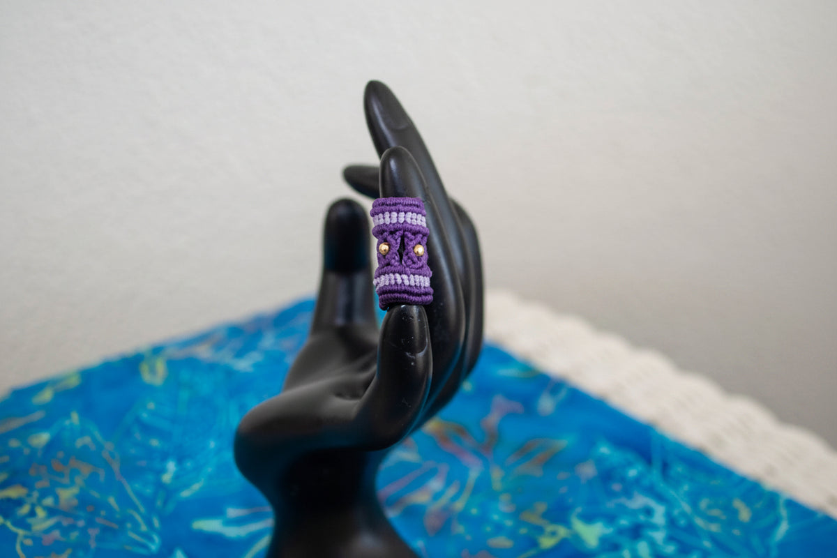 A purple and lavender dread bead with brass beads displayed on a black hand-shaped mannequin, set against a blue patterned fabric background.
