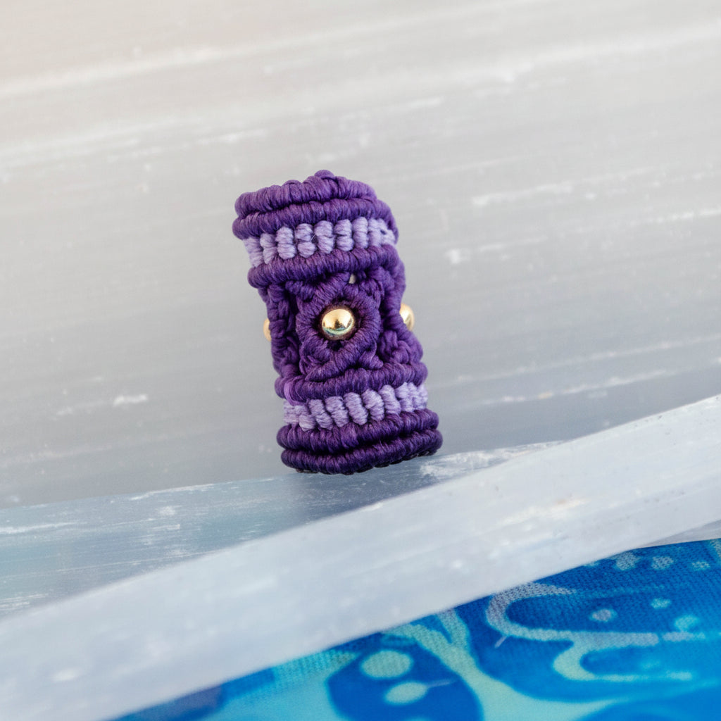 A purple and lavender macrame dread bead with a brass beads, photographed standing upright on a translucent clear surface against a pale gray background.