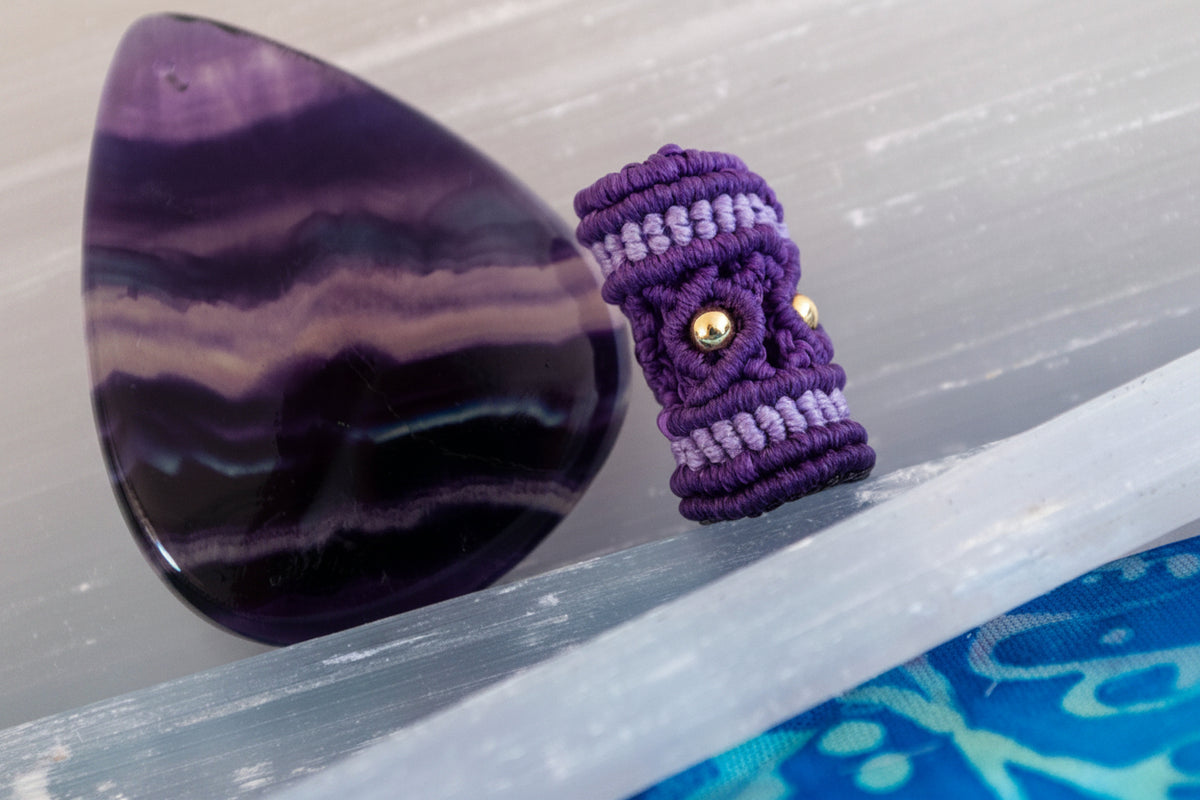 A purple and lavender macrame dread bead shaped with brass beads, displayed next to a polished purple agate stone on a light gray wooden surface.