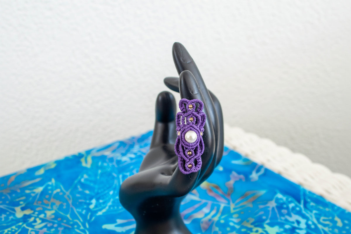 A handmade purple macrame dreadlock bead with an intricate lattice knotwork design, natural shell pearl accent, and gold seed beads, displayed on a black hand mannequin against a blue batik fabric background.