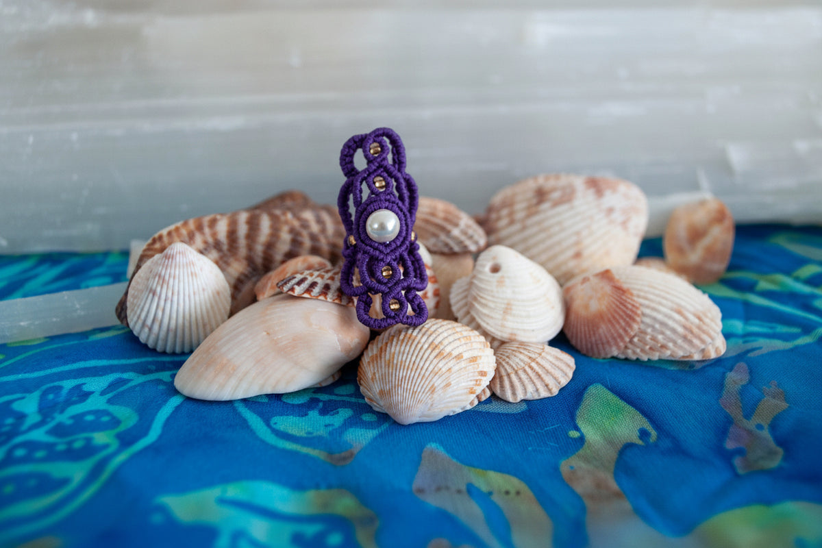 A handmade purple macrame dreadlock bead with an intricate lattice knotwork design, natural shell pearl accent, and gold seed beads, displayed upright among natural seashells on a blue batik fabric surface.