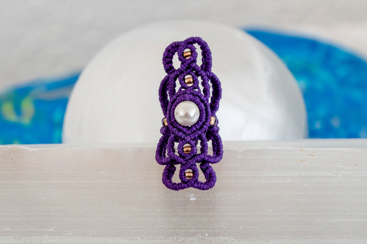 A handmade purple macrame dreadlock bead with an intricate lattice knotwork design, natural shell pearl accent, and gold seed beads, displayed upright on a white selenite crystal with a soft blue background.