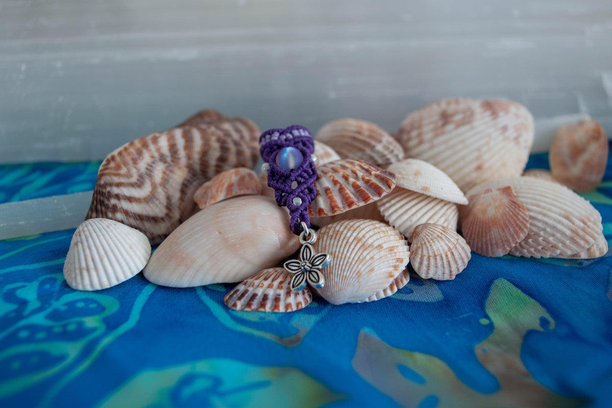 A handmade two-tone purple and lavender macrame dreadlock bead with a shimmering mermaid glass bead and a dangling silver-tone flower charm, nestled among natural seashells on a blue batik fabric surface