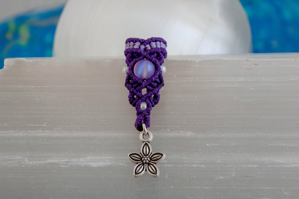 A handmade two-tone purple and lavender macrame dreadlock bead with a shimmering mermaid glass bead, pearlescent white seed beads, and a dangling silver-tone flower charm, displayed upright on a white selenite crystal with a soft blue background