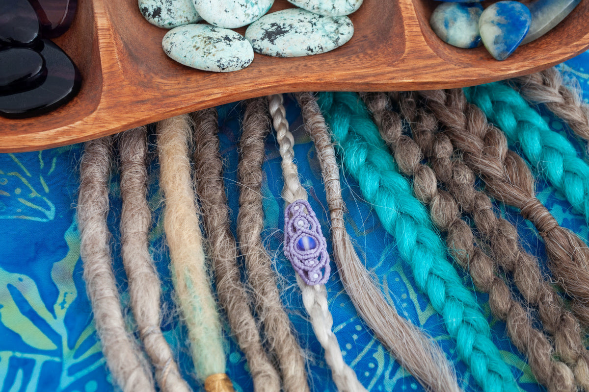 A close-up of a handmade lavender macrame dreadlock bead with lattice knotwork and a shimmering iridescent mermaid glass bead, worn on a natural dreadlock alongside various loc styles on a blue batik fabric surface beneath a wooden crystal tray. Only the lavender lattice dreadlock bead is for purchase.