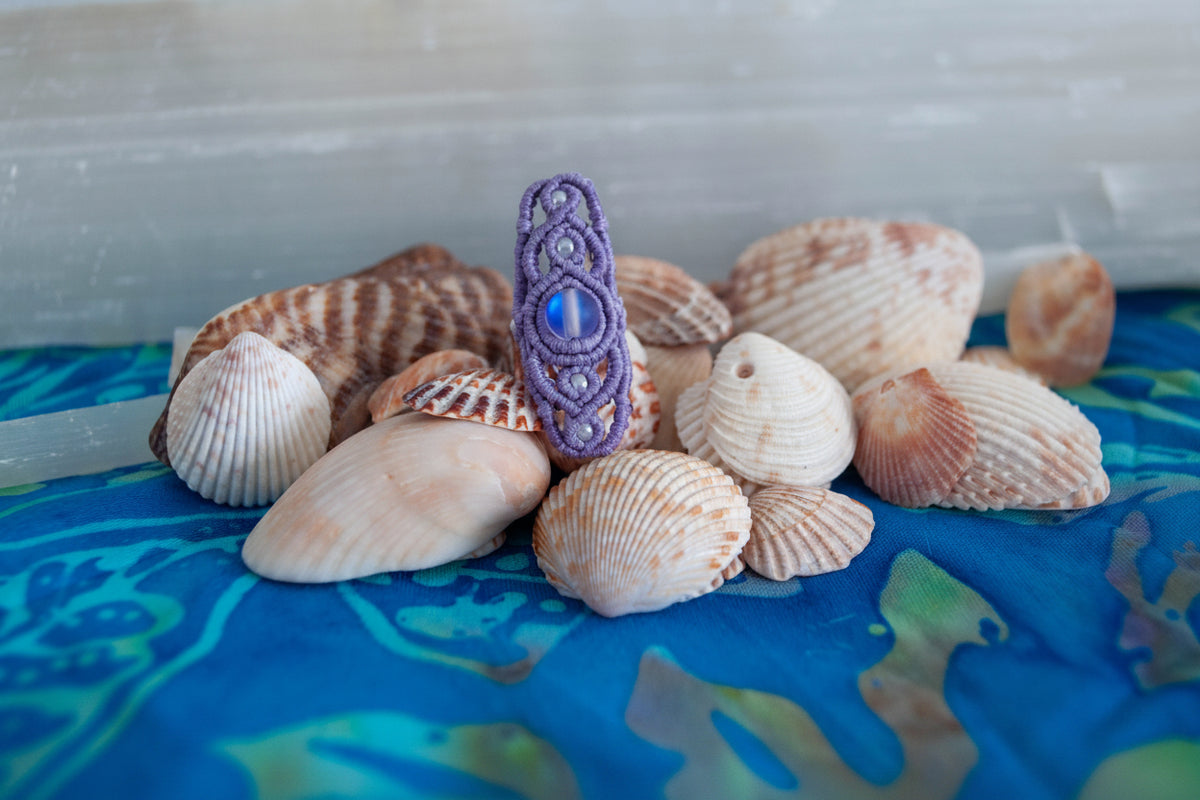 A handmade lavender macrame dreadlock bead with intricate lattice knotwork, a shimmering iridescent mermaid glass bead, and pearlescent white seed beads, displayed upright among natural seashells on a blue batik fabric surface