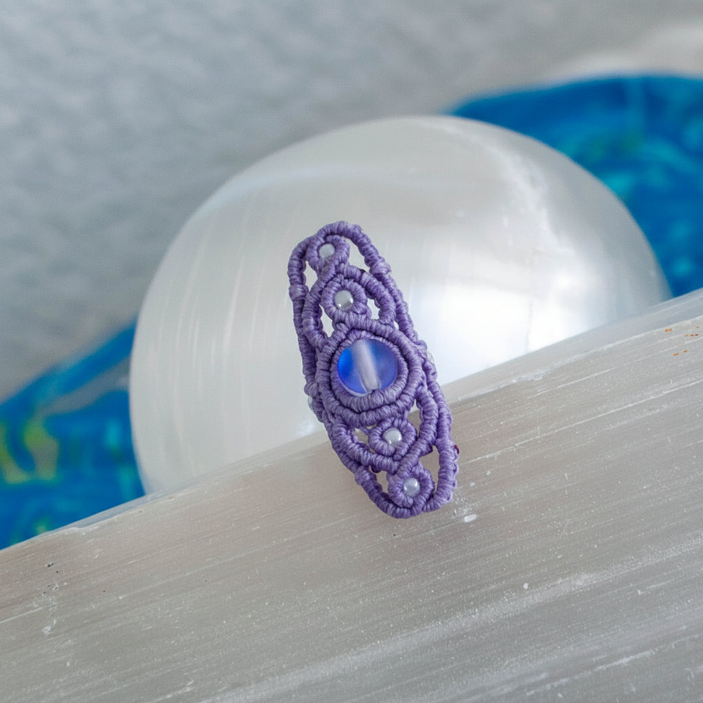 A handmade lavender macrame dreadlock bead with intricate lattice knotwork, a shimmering iridescent mermaid glass bead, and pearlescent white seed beads, displayed on a white selenite crystal with a soft blue background