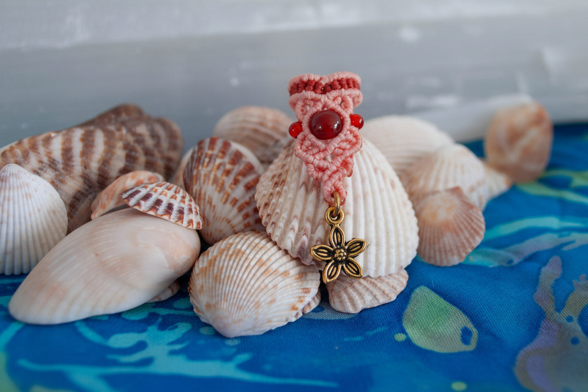 A handmade two-tone coral and rust macrame dreadlock bead with drop knotwork, a deep red carnelian bead accent, and a dangling brass flower charm, displayed upright among natural seashells on a blue batik fabric surface.