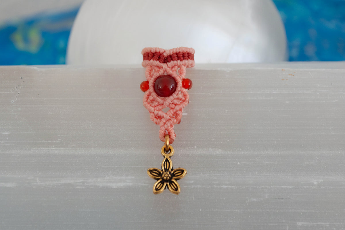A handmade two-tone coral and rust macrame dreadlock bead with drop knotwork, a deep red carnelian bead accent, carnelian side beads, and a dangling brass flower charm, displayed upright on a white selenite crystal with a soft blue background.