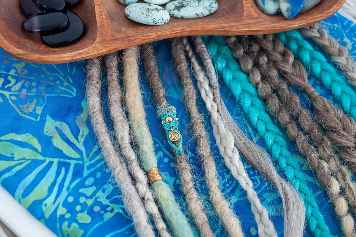 A close-up of a handmade two-tone teal and brown macrame dreadlock bead with a brass accent bead and brass spiral charm, worn on a natural dreadlock alongside various loc styles on a blue batik fabric surface beneath a wooden crystal tray. Only the teal dreadlock bead is for purchase.