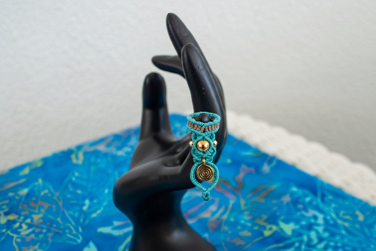 A handmade two-tone teal and brown macrame dreadlock bead with a brass accent bead and a dangling brass spiral charm, displayed on a black hand mannequin against a blue batik fabric background