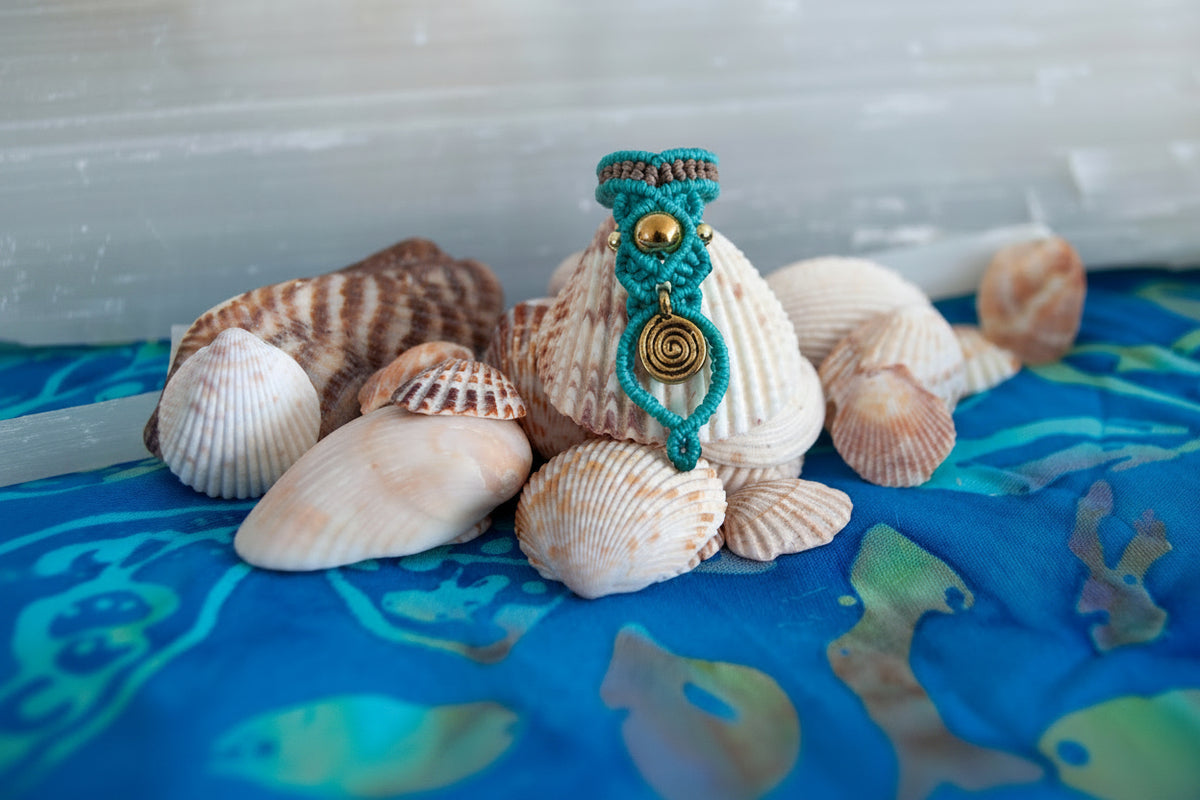 A handmade two-tone teal and brown macrame dreadlock bead with a brass accent bead and a dangling brass spiral charm, displayed upright among natural seashells on a blue batik fabric surface.