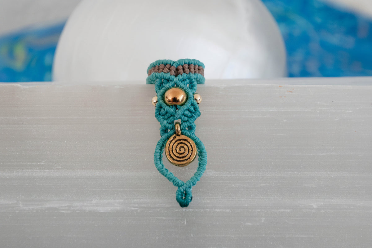 A handmade two-tone teal and brown macrame dreadlock bead with a brass accent bead and a dangling brass spiral charm, displayed upright on a white selenite crystal with a soft blue background.