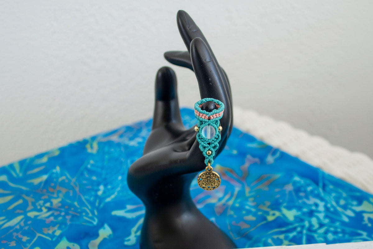 A handmade two-tone teal and peach macrame dreadlock bead with a shimmering mermaid glass bead, round brass beads, and a dangling brass sand dollar charm, displayed on a black hand mannequin against a blue batik fabric background