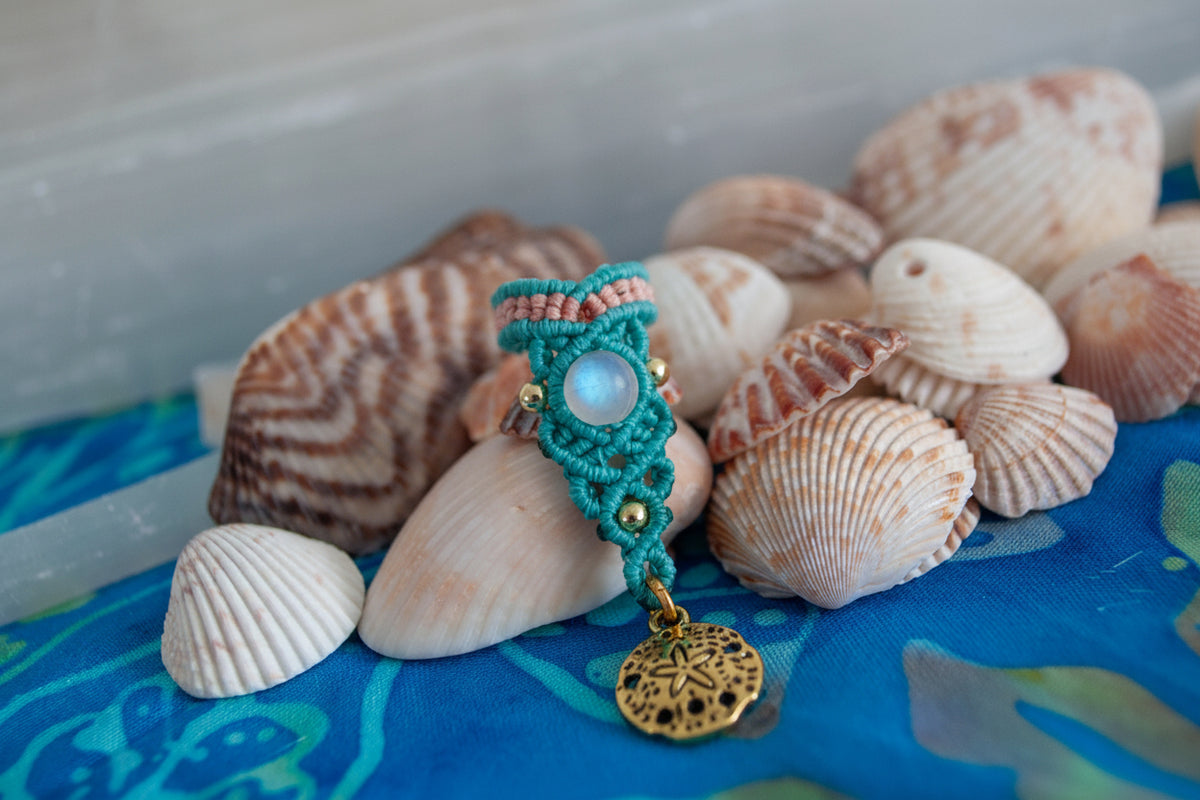 A handmade two-tone teal and peach macrame dreadlock bead with a shimmering mermaid glass bead and a dangling brass sand dollar charm, nestled among natural seashells on a blue batik fabric surface