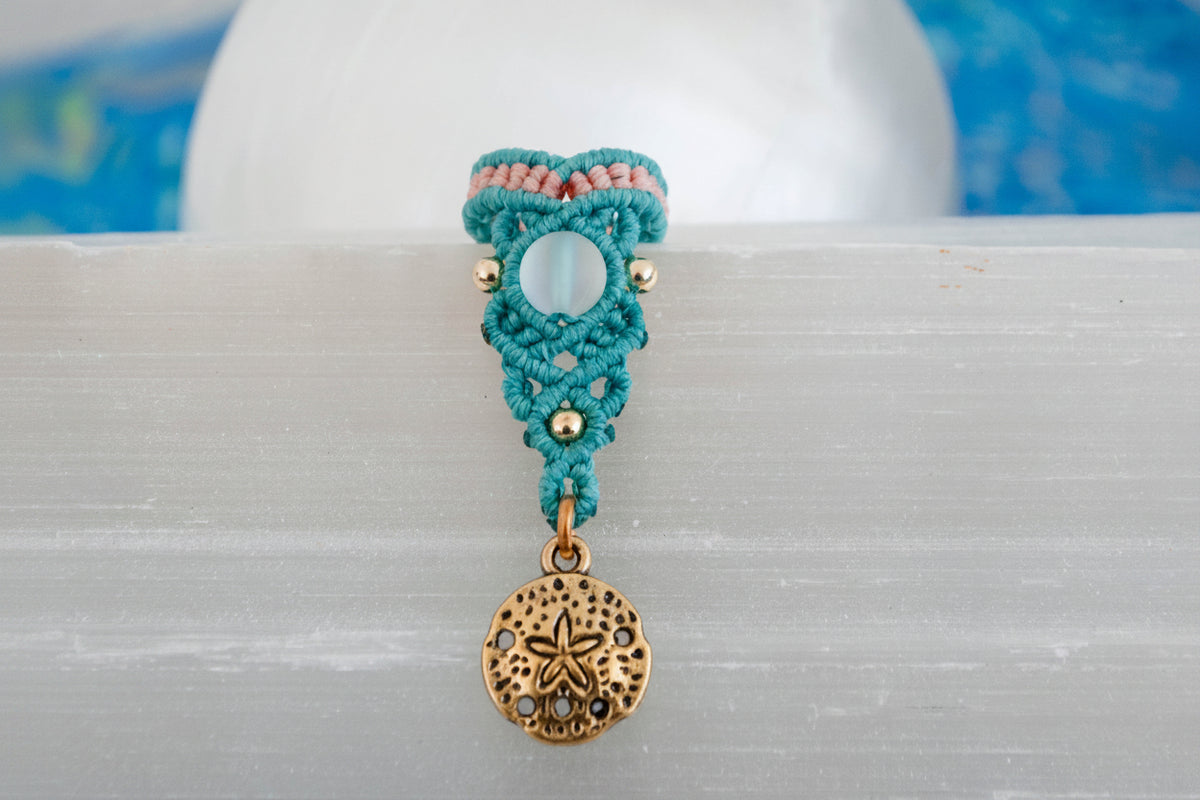 A handmade two-tone teal and peach macrame dreadlock bead with a shimmering mermaid glass bead, round brass beads, and a dangling brass sand dollar charm, displayed upright on a white selenite crystal with a blue batik fabric background